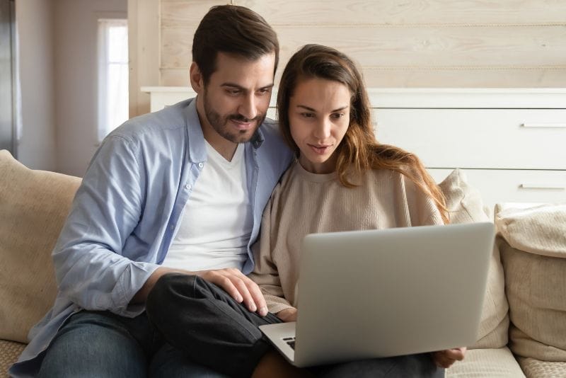 Mężczyzna i kobieta siedzą blisko siebie na kanapie, patrząc na ekran laptopa i uśmiechając się. Wyglądają na skupionych i zaangażowanych, w przytulnym, dobrze oświetlonym salonie.
