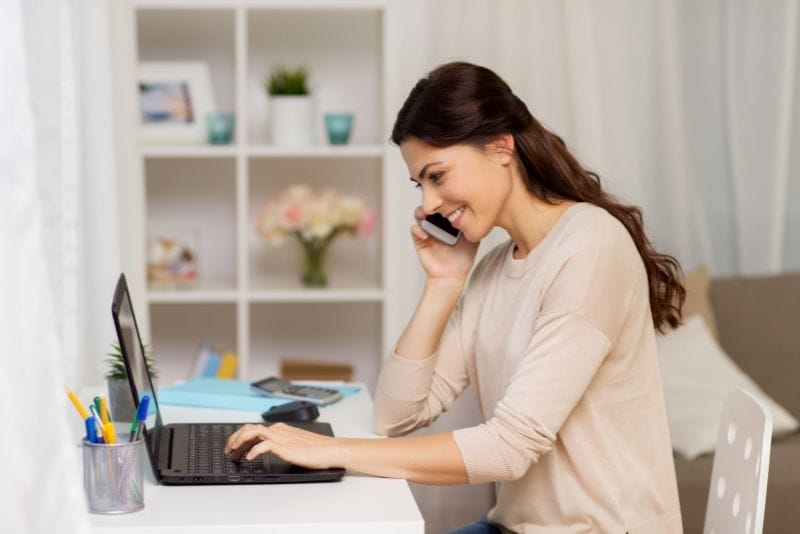 Kobieta siedzi przy biurku, pracuje na laptopie i rozmawia przez smartfon, umawiając wizytę u terapeuty. Na biurku leżą artykuły biurowe, notatnik i kubek z długopisami, a w tle widać półki i wazon z kwiatami.