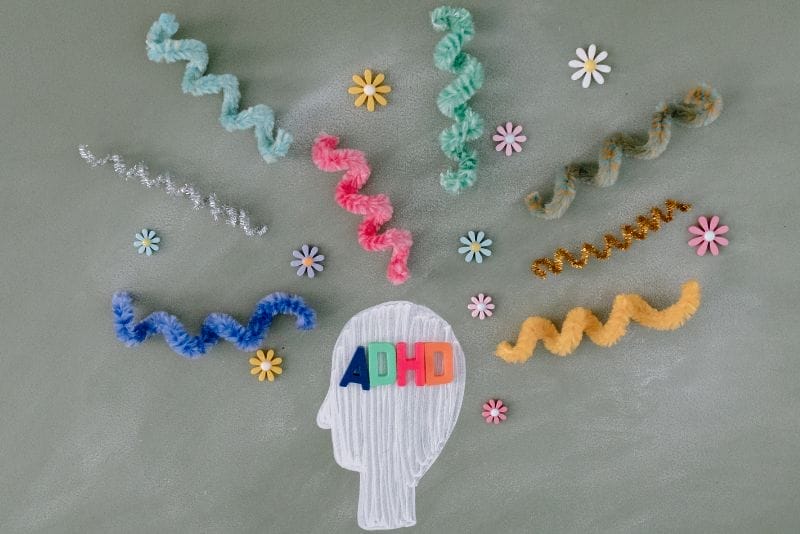 Rysunek głowy oznaczonej ADHD z kolorowymi czyścikami do rur i papierowymi kwiatami rozchodzącymi się wokół niej, reprezentującymi rozproszone myśli na szarym tle.