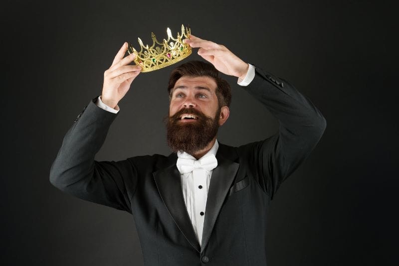 Mężczyzna w eleganckim garniturze trzyma złotą koronę nad głową, uśmiechając się i patrząc w górę, na tle czarnego tła.