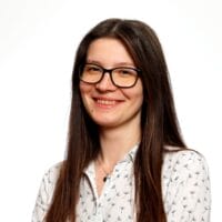 Kobieta w okularach. uśmiechnięta, w eleganckiej bluzce, siedzi na krześle ma specione dłonie