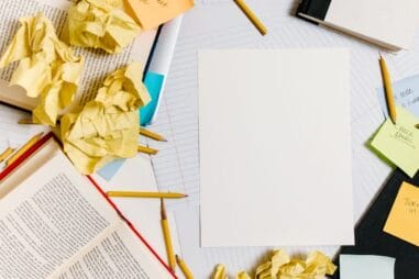 Pusta kartka papieru otoczona zmiętymi żółtymi papierami, ołówkami, otwartymi książkami i karteczkami samoprzylepnymi na zagraconym biurku, sugerująca scenę burzy mózgów lub blokady pisarskiej.