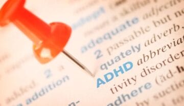 Zbliżenie strony słownika z terminem ADHD napisanym niebieskim tekstem, wyróżnionym czerwoną pinezką wskazującą na niego. Otaczający tekst jest lekko rozmyty.