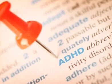 Zbliżenie strony słownika z terminem ADHD napisanym niebieskim tekstem, wyróżnionym czerwoną pinezką wskazującą na niego. Otaczający tekst jest lekko rozmyty.