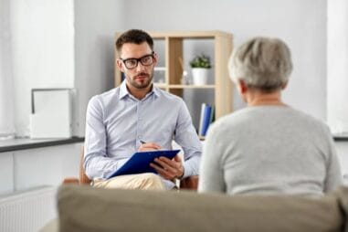 Mężczyzna w okularach trzymający schowek uważnie słucha starszej kobiety z krótkimi siwymi włosami podczas sesji doradczej lub terapeutycznej w jasnym biurze.