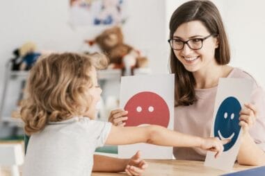 Kobieta w okularach trzyma dwie papierowe twarze, jedną czerwoną i smutną, jedną niebieską i szczęśliwą, podczas gdy małe dziecko wskazuje na niebieską, szczęśliwą twarz. Siedzą przy stole w jasno oświetlonym pokoju.