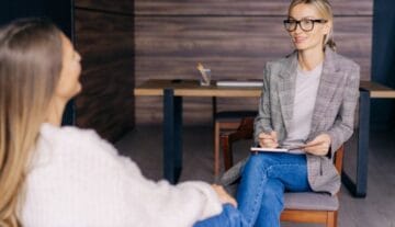 Szczecińska psychoterapeutka w okularach i kraciastej marynarce siedzi z notesem, uśmiecha się i rozmawia z inną kobietą w przytulnym swetrze w biurze, w tle stoi drewniane biurko.