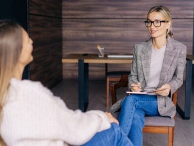 Szczecińska psychoterapeutka w okularach i kraciastej marynarce siedzi z notesem, uśmiecha się i rozmawia z inną kobietą w przytulnym swetrze w biurze, w tle stoi drewniane biurko.