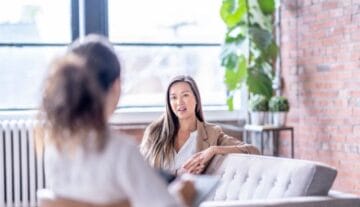 Dwie kobiety rozmawiają w jasnym, nowoczesnym centrum psychoterapii w Szczecinie z ceglanymi ścianami i dużymi oknami. Jedna, prawdopodobnie psychoterapeutka ze Szczecina, siedzi na kanapie i mówi, podczas gdy druga uważnie słucha i robi notatki.