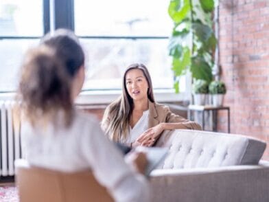 Dwie kobiety rozmawiają w jasnym, nowoczesnym centrum psychoterapii w Szczecinie z ceglanymi ścianami i dużymi oknami. Jedna, prawdopodobnie psychoterapeutka ze Szczecina, siedzi na kanapie i mówi, podczas gdy druga uważnie słucha i robi notatki.