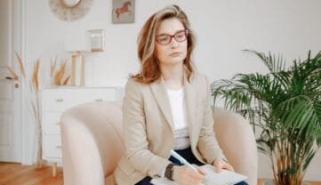 Terapeuta uzależnień ze Szczecina, w okularach i beżowej marynarce, siedzi w jasnym fotelu, pisząc w notesie. Wydaje się skupiona. W pokoju znajduje się roślina, sztuka ścienna i minimalistyczny wystrój.