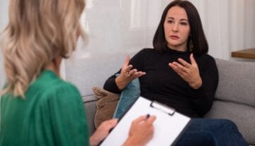 Kobieta o ciemnych włosach siedzi na kanapie, gestykulując rękami, gdy rozmawia z inną kobietą trzymającą kartkę i długopis, sugerując sesję terapeutyczną lub doradczą.