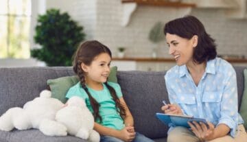 Kobieta z notatnikiem rozmawia z uśmiechniętą dziewczynką o długich warkoczach siedzącą na kanapie z pluszowym misiem.