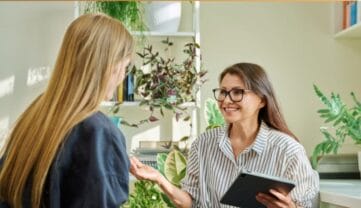 Kobieta z długimi, prosto ułożonymi włosami rozmawia z drugą kobietą o brązowych włosach w okularach, która trzyma tablet. Obie siedzą w jasnym pomieszczeniu z roślinami doniczkowymi w tle.