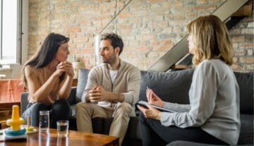 Para siedzi na kanapie, rozmawia poważnie i trzyma się za ręce, podczas gdy szczeciński psychoterapeuta uważnie słucha i robi notatki podczas sesji terapeutycznej w nowoczesnym pokoju z ceglanymi ścianami.