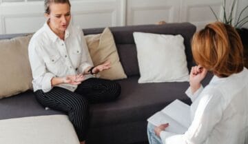 Kobieta w jasnej koszuli opowiada gestykulując na kanapie, podczas gdy druga kobieta w białej bluzie, notując w zeszycie, uważnie jej słucha w przytulnym pomieszczeniu.