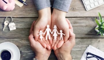 Dwie dorosłe ręce delikatnie trzymają mniejsze dłonie, które kołyszą papierową wycinankę czteroosobowej rodziny. Scena rozgrywa się na drewnianym stole, obok którego znajdują się filiżanka do kawy, klucze, roślina, klawiatura i skarbonka.
